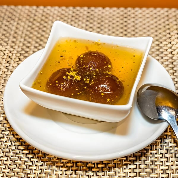 Three gulab jamun in a white bowl with yellow syrup and a spoon on a woven placemat.