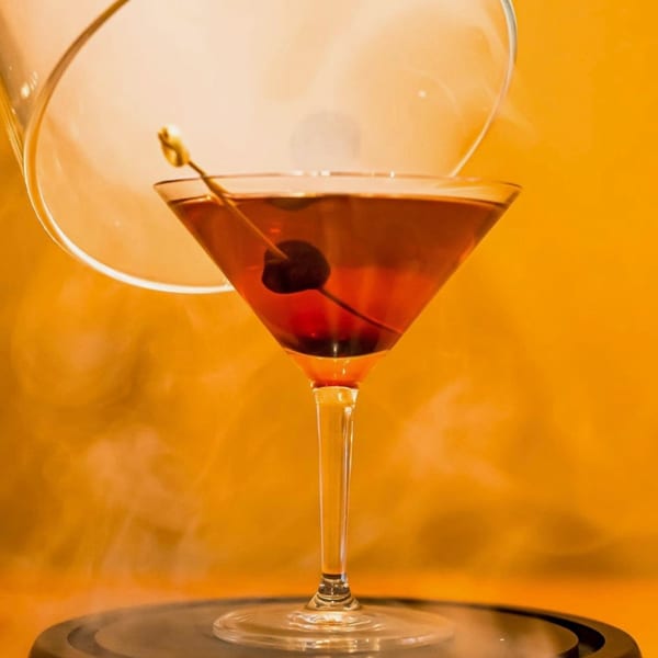 A smoked cocktail with a cherry garnish in a martini glass, against an orange background.