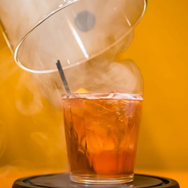 Smoky orange cocktail with ice and a straw, served under a glass cloche.
