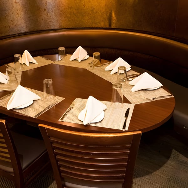 An oval dining table set with placemats, folded napkins, and glasses, surrounded by a dark booth.